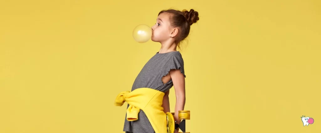 Žvýkající a žlutou bublinu ze žvýkačky nafukující mladá slečna držící za zády skateboard oblečena v šedém tričku s ovázanou žlutou mikinou se žlutým pozadím | Pro článek: