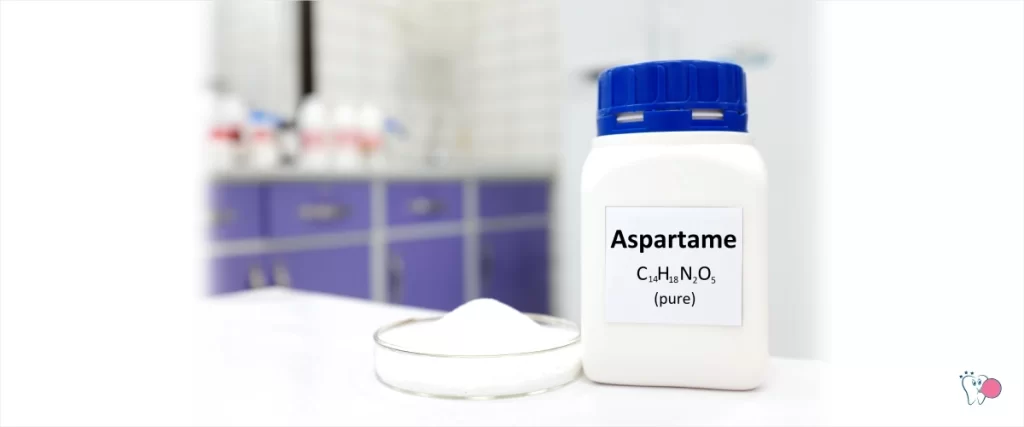 Na fotografii je znázorněno umělé sladidlo aspartam jak v podobě bílého cukru v ploché kulaté transparentní laboratorní misce tak v bílé neprůhledné láhvi s modrým víčkem a černým popisem "aspartam" a chemickým vzorcem v pozadí laboratoř | pro článek - Aspartam je karcinogenní | pro web: zdravezvykacky.cz | Zdroj: shutterstock