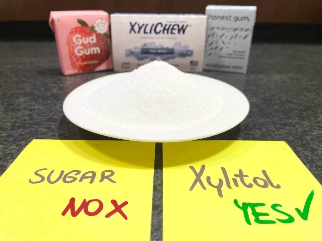 In the photo in the foreground on a yellow piece of paper on the left side the word Sugar and the red English disagreement NO with a red cross and on the right side the word Xylitol with the green English text YES and a green whistle in the middle on a plate a pile of sugar and in the background healthy gum with xylitol from Gud brands Gum, Xylichew and honest gum | For the article: Why you need xylitol in your gum | For the website: healthchewinggum.com | Author and graphic editing: Jiří Samuel