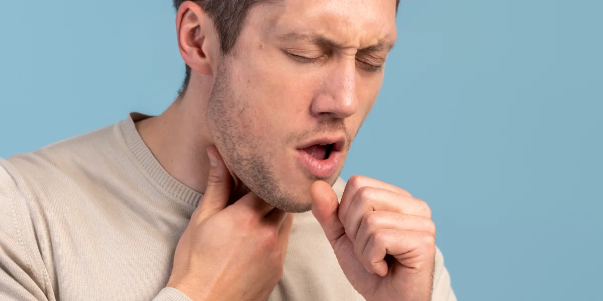 A young man holding his neck with his hand and coughing, with a sore throat and dry mouth, his eyes closed and his hand clenched into a fist to cover his cough, in a home setting | Image source: shutterstock.com | For the article: What causes dry mouth and how to relieve it | For the website: healthchewinggum.com